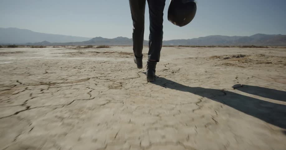 A person walks through the desert in leather clothing holding a motorcycle helmet