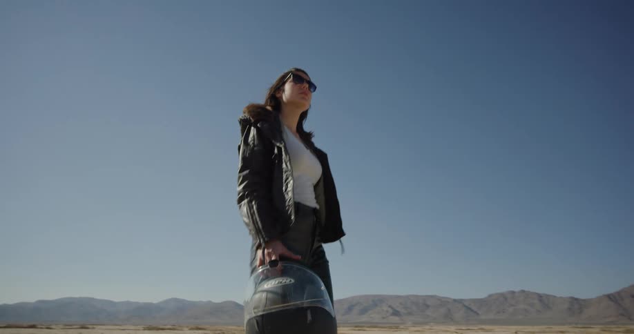 A woman in leather jacket and pants walking through a desert carrying a motorcycle helmet