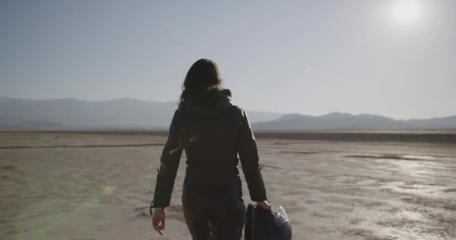 Woman walks across arid cracked dirt toward road and mountains