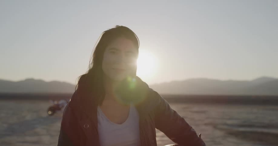 Female motorcyclist stands and smiles at the camera on the dry plains at sunset