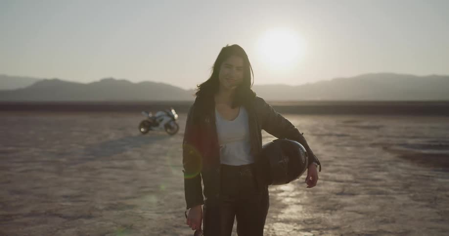 A woman in leather standing in the desert with her motorcycle near a road