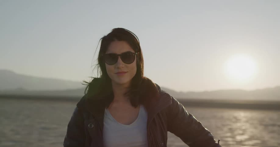 Female motorcyclist takes off her sunglasses and looks at the camera with a smile on a dry plain at sunset