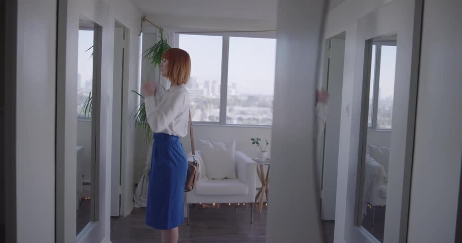 A woman stopping before a mirror to adjust her hair and skirt before continuing down a hallway