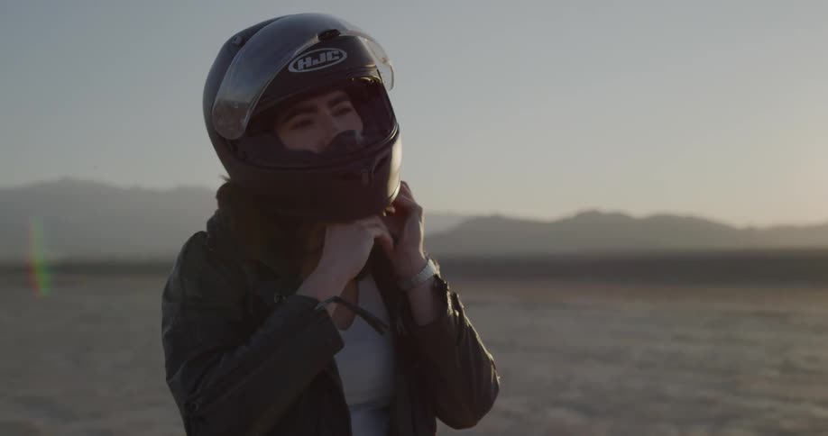 Female motorcyclist gets on the motorcycle on the dry plain at sunset