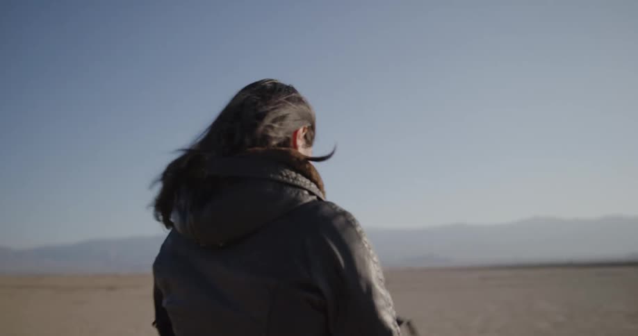 A woman in a desert walking towards a motorcycle and putting on a helmet