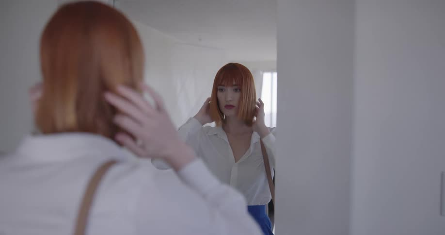 Red haired woman looks in the mirror and checks appearance before going out at home