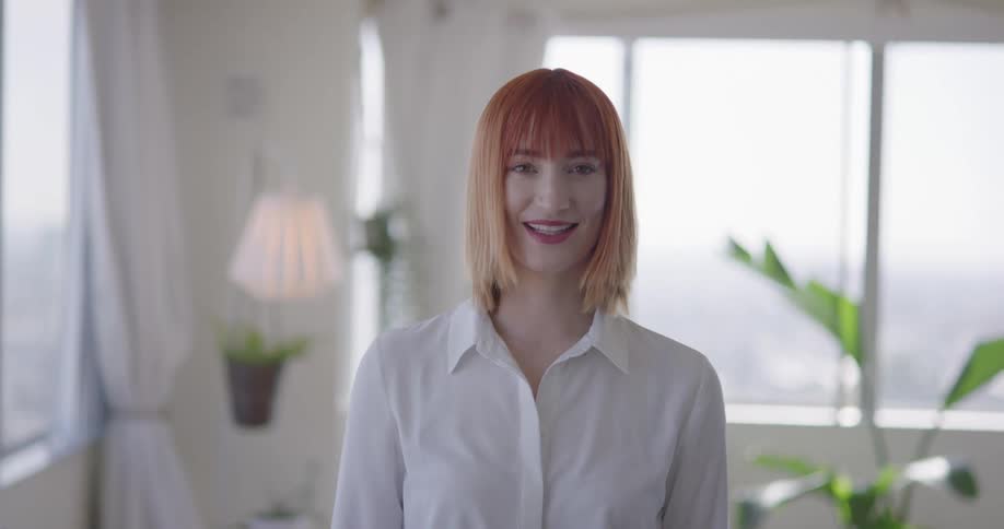 A redheaded woman smiles as she stands in her living room with crisp white walls and blooming plants