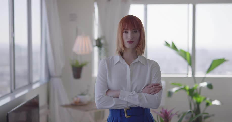 A woman standing in a room overlooking a city while looking into the camera with crossed arms
