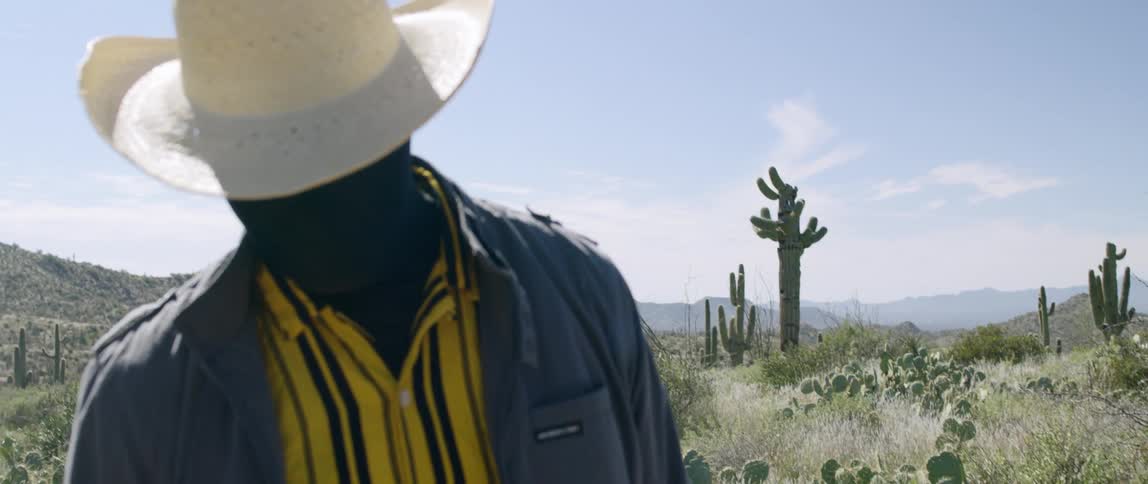 A mans face is covered in black fabric as he poses standing in the desert with his head down