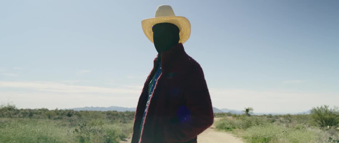 A black man in a cowboy hat turning around to face the camera while standing in a dirt road through a grassland