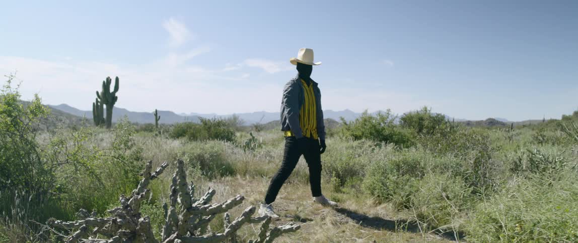 A man wearing a black full head and face cover stands up from a crouched position in the desert