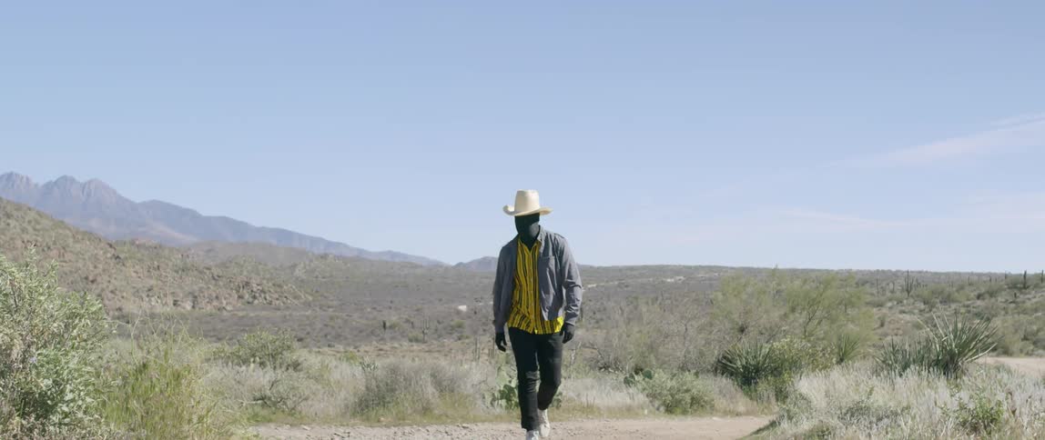 Man wearing black face mask and cowboy hat walks on a path on the desert