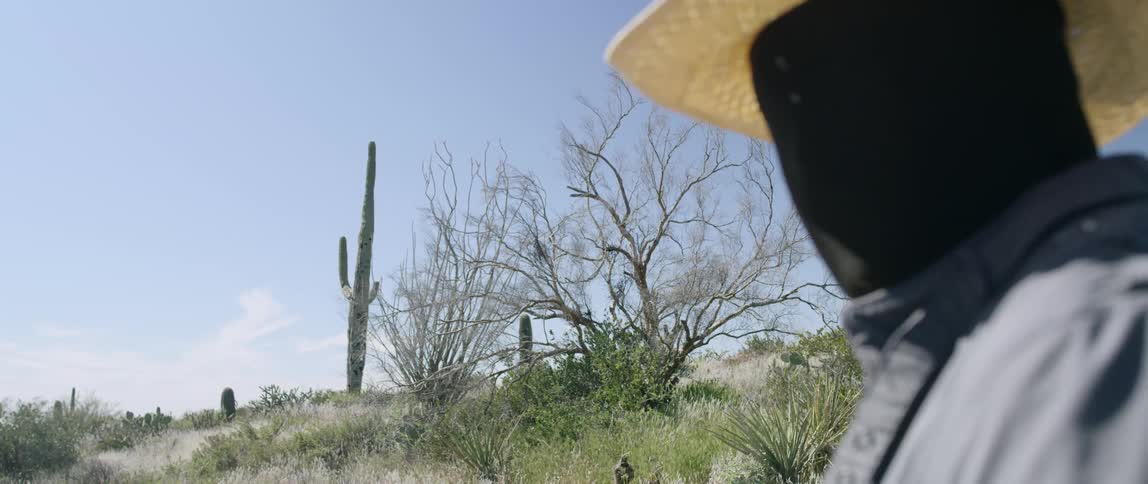 A man wearing a black full face and head cover under a straw hat stands in the desert near cactus and brush