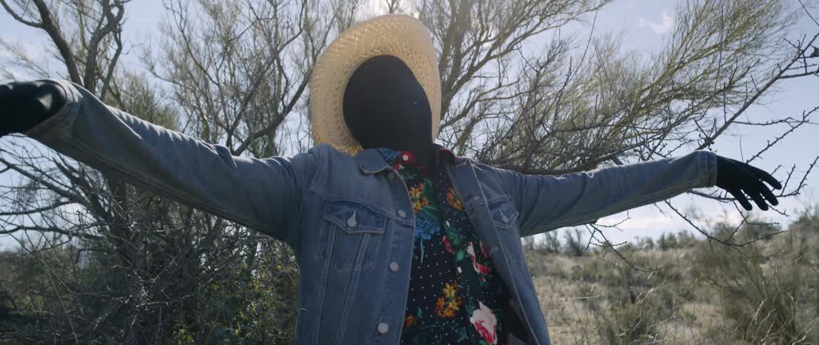 A man with his face completely covered in black fabric raising both arms wide while standing outdoors in a desert