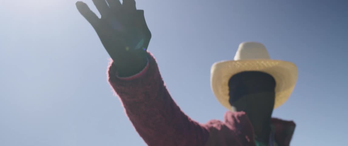 A man wearing a full black face covering and black gloves poses with his arm outstretched while standing in the desert