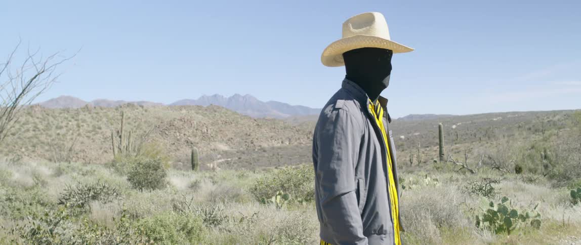 Man wearing black face mask and cowboy hat stands on desert