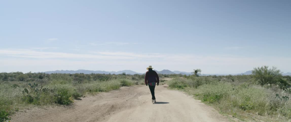 Cowboy walks down dirt road between scrub covered plains toward mountains on the horizon