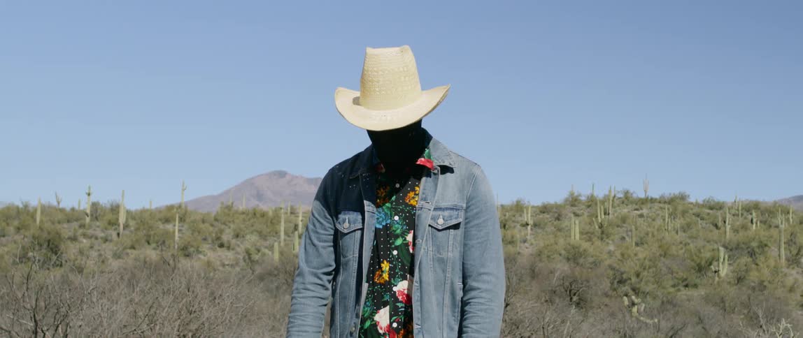 A man wears a black mask that completely covers his skin as he poses in the desert
