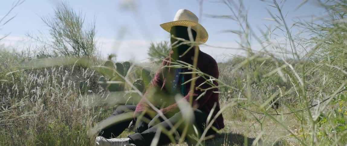 A cowboy has his face and hands completely covered in black fabric as he sits in a field