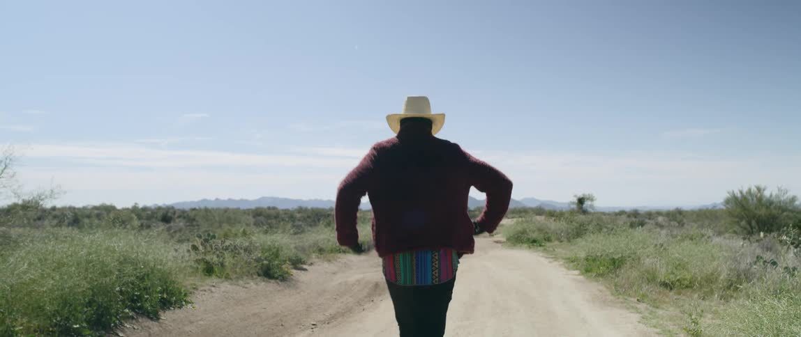 A man with a covered face wearing a cowboy hat walking down a dirt road through a field