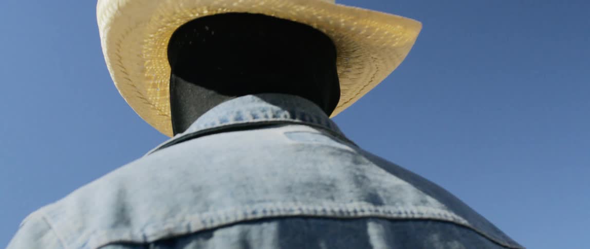 Cowboy wearing black full face and head covering stands under blue sky