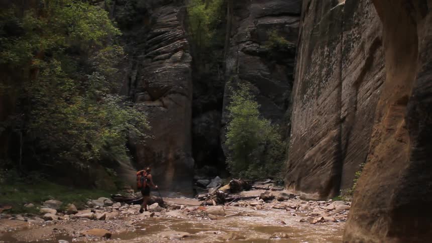 A hiker walking alongside a shallow creek that is surrounded by cliffs and trees