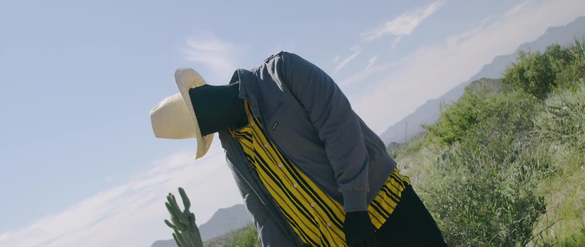 A man with his face and hands covered in black fabric standing with an odd posture in a desert