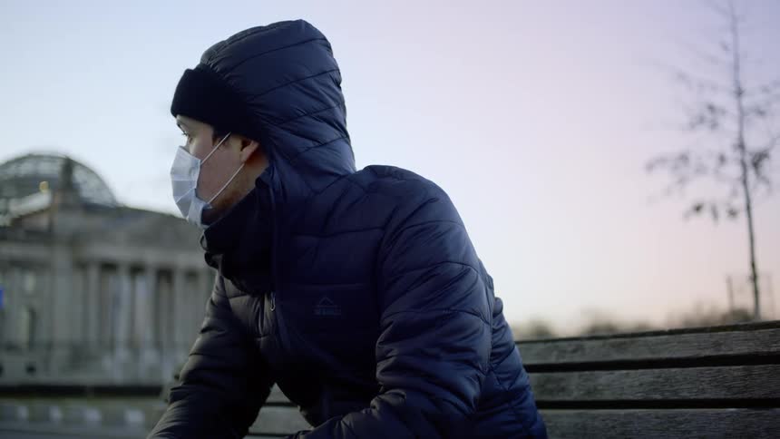 A man in a face mask sitting outdoors on a wooden park bench