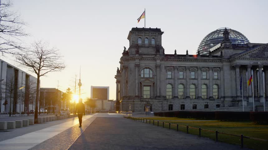 A person walks down an empty sidewalk past a large building with flags flying in front and an american flag on top