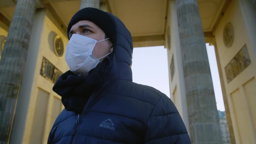 A man standing outside with a mask and a coat with pillars in the background