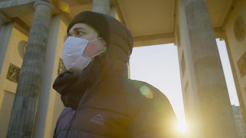 A person with a mask and a coat standing in front of pillars
