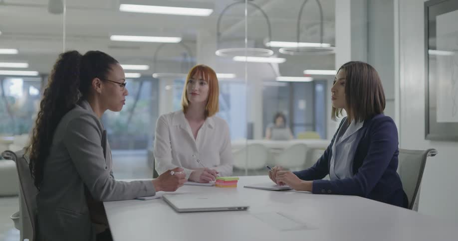 Three women sit in a conference room and talk while one takes notes