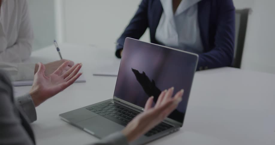 A woman gestures with her hands in front of her laptop while meeting with two other women in a conference room