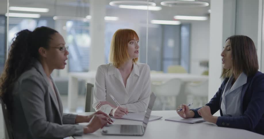 Three woman sitting at a desk in an office talking and writing