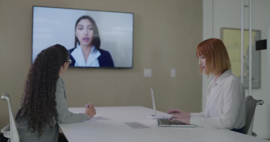 Two women sit in a conference room and talk with another woman on a video call