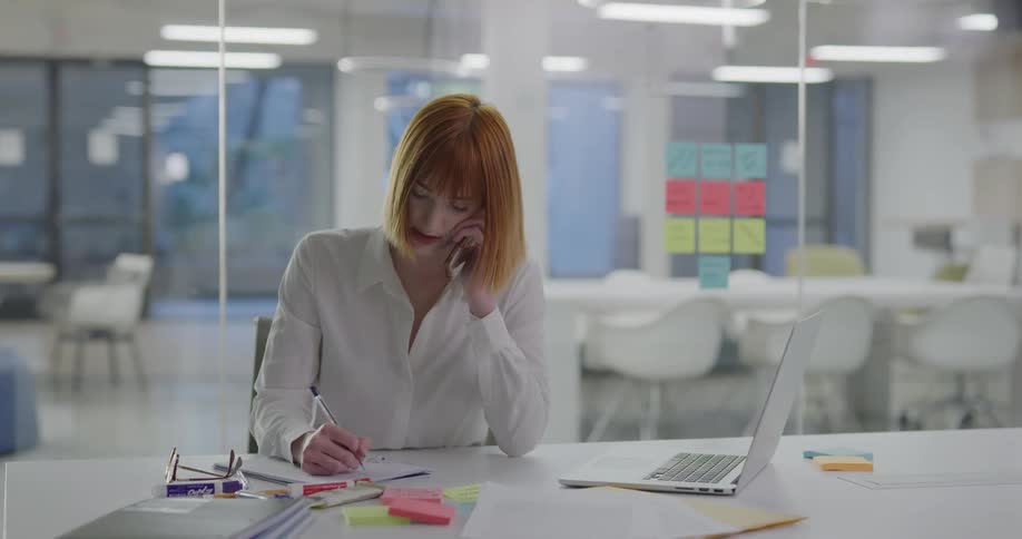 A woman sitting at a desk talking on the phone and writing on a notepad