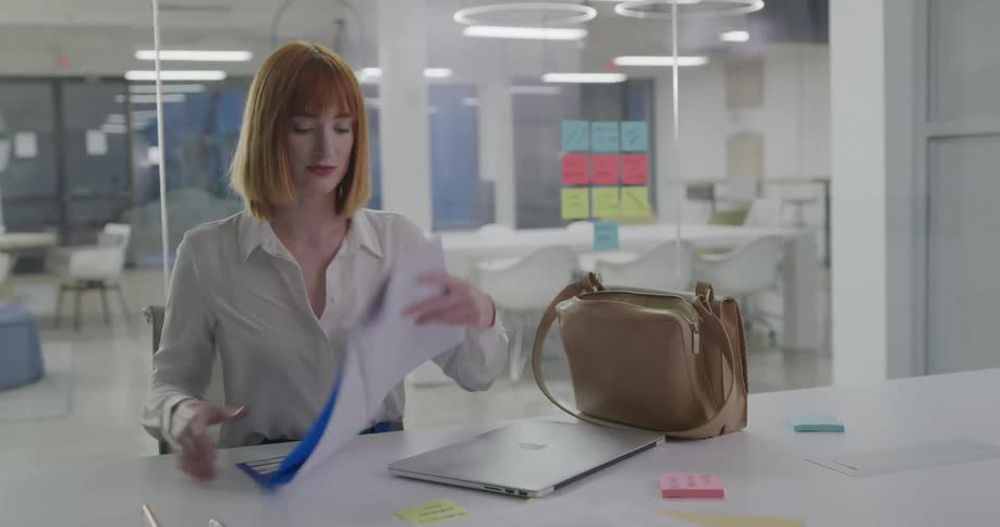 A woman in a conference room shuts down her laptop and packs her files in a bag then gets up and carries everything out