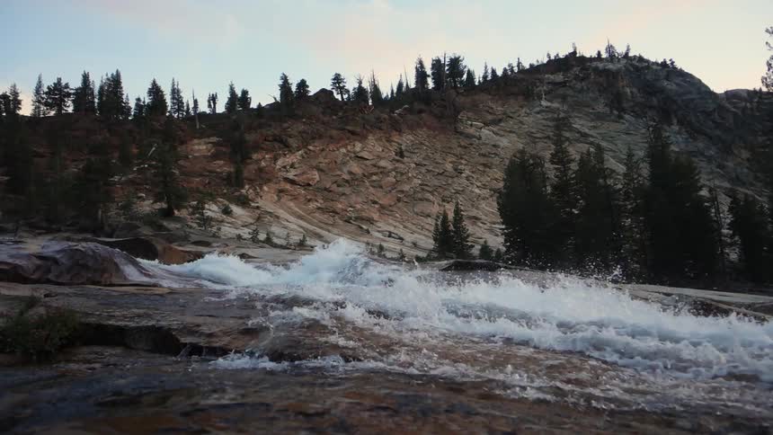 Water rushing quickly over a rocky surface with a hill and pine trees in the background