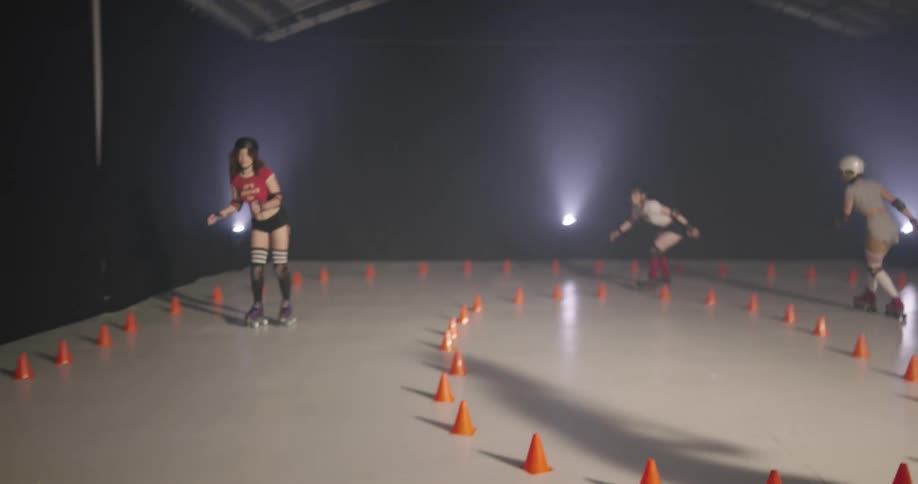 Four women in shorts helmets and pads roller skate around a small rink with a track marked by orange cones
