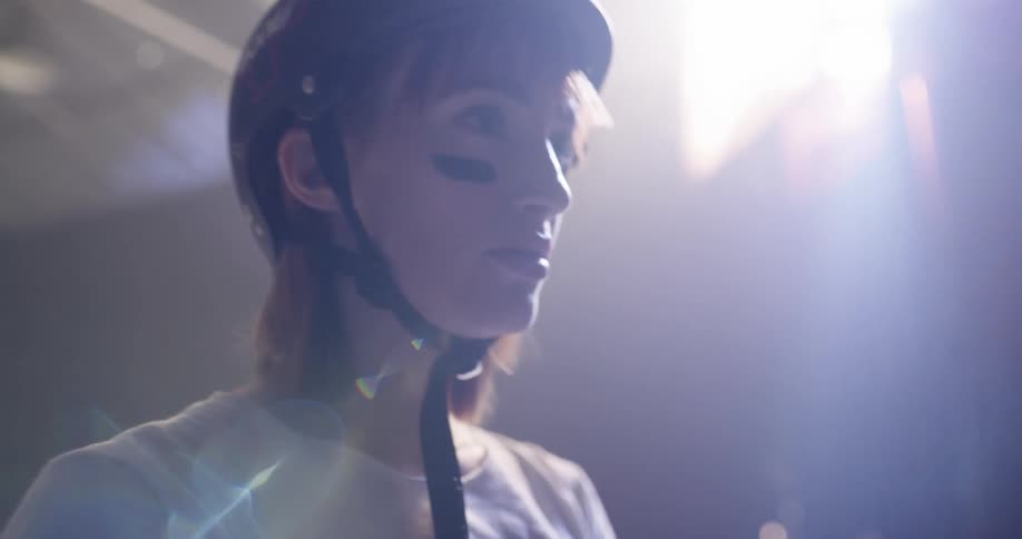 A woman wearing a helmet wrist guards and eye black drinks from a bottle of water in an arena with lights shining