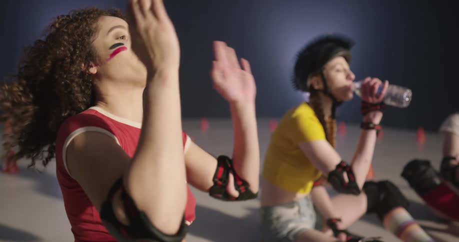 Three women with roller skates on sit on the floor and drink water and take off a helmet