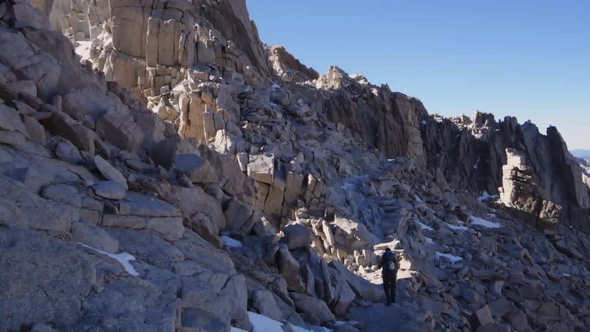 A person using hiking poles ascends a path in a rocky landscape with patches of snow