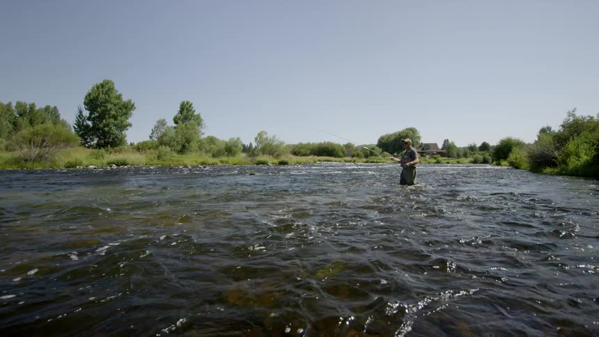 A fisherman stands in a large body of moving water holding his fishing pole with a bright blue sky behind him