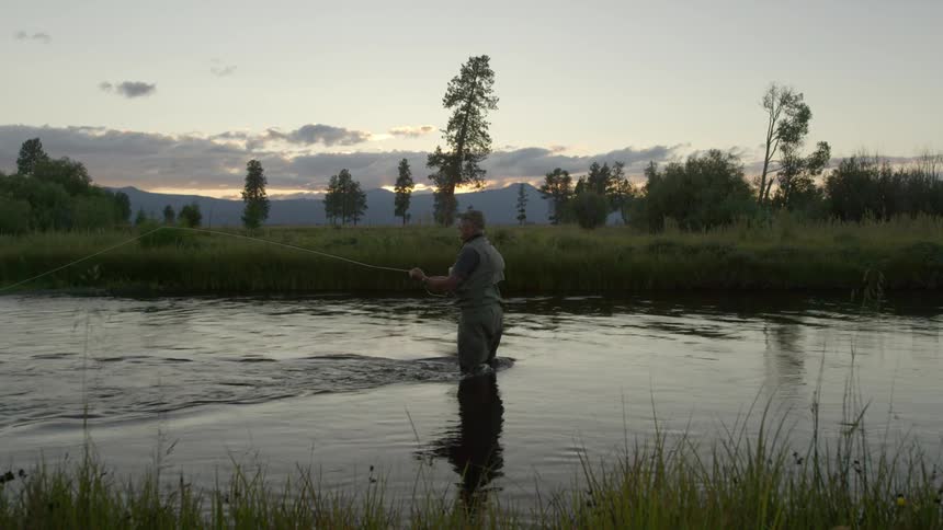 A fisherman stands in a flowing body of water surrounded by grass and trees fishing during sunset