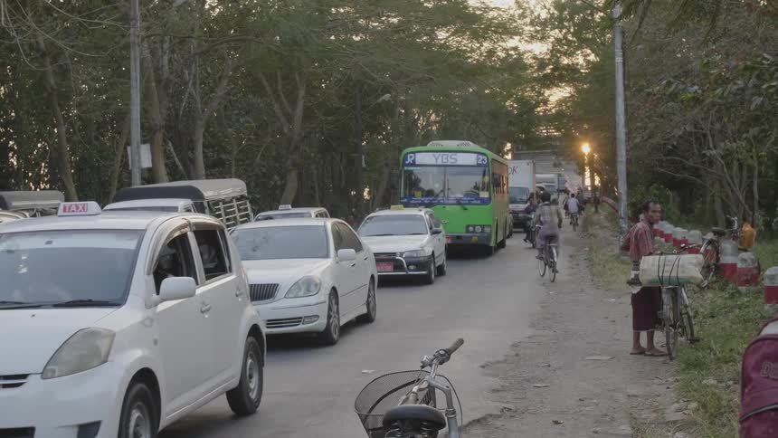 A man with a load on the back of his bicycle stands on the dirt road side while heavy traffic travels on the street beside him