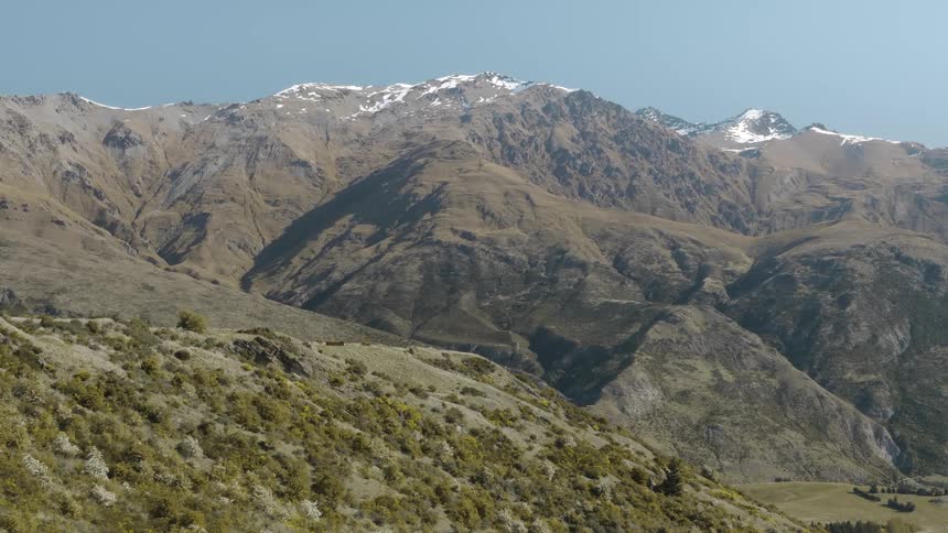 A snow capped mountain range above a hill covered in shrubs on a sunny day