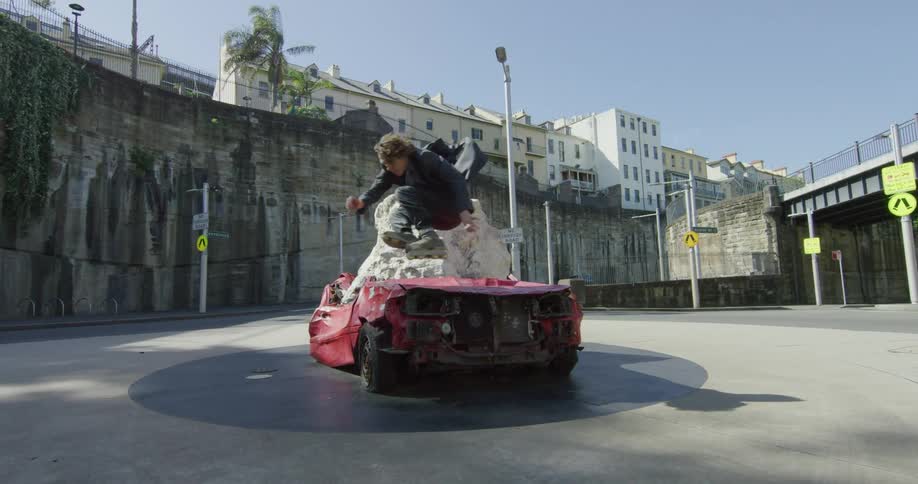 A teen roller skates and jumps over a smashed up car
