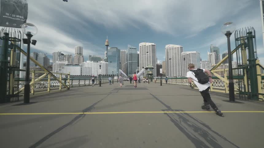 A teenager roller skates across a busy bridge with many people walking