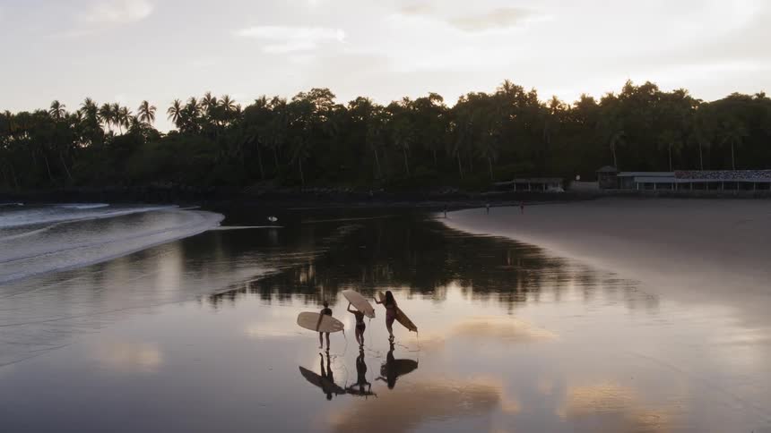 Three surfers carry surfboards on a sandy beach at sunset