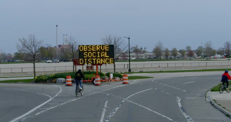 Two bicyclist pass past a sign about social distancing near a fenced parking lot
